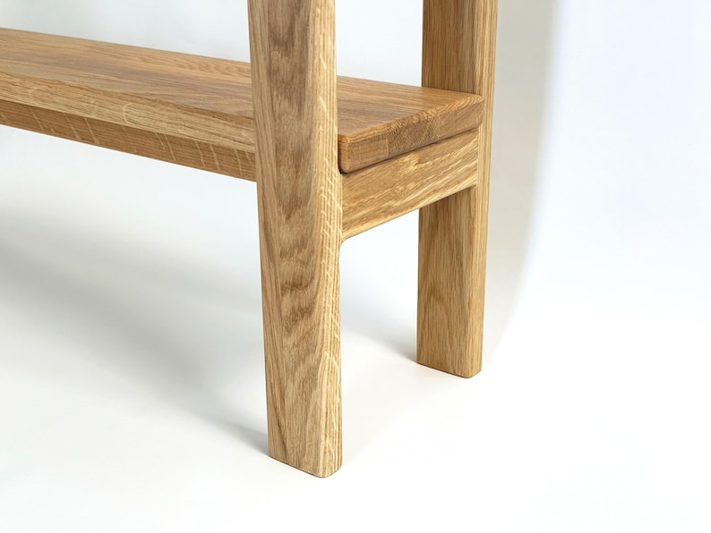 May include: A close-up of a wooden shelf with a light brown finish. The shelf has a rectangular design with a lower shelf and two supporting legs. The wood grain is visible, and the piece is set against a white background.