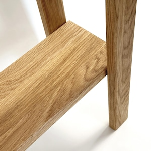 May include: Close-up of a section of a wooden table or shelf. The wood has a natural, light brown color with visible grain patterns. The design features a shelf supported by a leg, showcasing the joinery and craftsmanship.