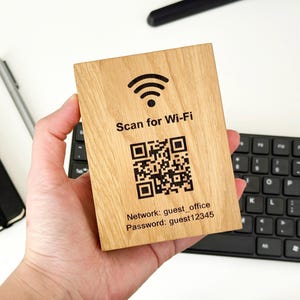 May include: A rectangular wooden sign with the text "Scan for Wi-Fi" and a QR code. Below the QR code are the network name and password. The sign is held in a hand.