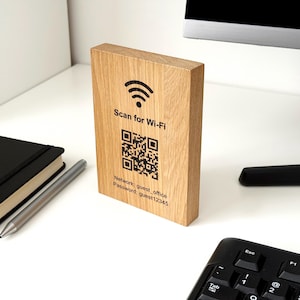 Könnte beinhalten: Ein hölzernes WLAN-Schild mit einem QR-Code und dem Text "Scan for Wi-Fi". Das Schild zeigt auch den Netzwerknamen und das Passwort an. Ein schwarzes Notizbuch, ein silberner Stift und eine Tastatur sind ebenfalls sichtbar.