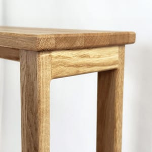May include: Close-up of a light brown wooden table. The table has a rectangular top and four square legs. The wood grain is visible, and the table appears to be made of solid wood. The table is a simple, modern design.