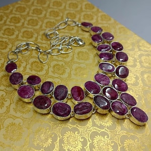 May include: A silver necklace with oval, deep red gemstones. The gemstones are set in individual bezels and linked with a silver chain. The necklace is displayed on a gold-coloured surface.