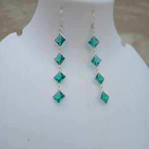 May include: A pair of dangling earrings with a silver hook and four emerald-green, square-cut gemstones. The gemstones are set in silver and cascade down the length of the earrings. The earrings are displayed on a white mannequin.
