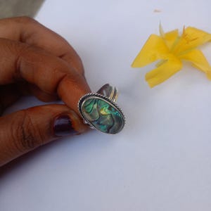 May include: A silver ring featuring an oval-shaped abalone shell inlay with iridescent green, blue, and gold tones. The setting is framed by a twisted rope-style metal border, and the band has a smooth, polished finish on the sides.