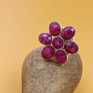 Op de afbeelding: Een ring met een bloemvormige cluster van zes gefacetteerde, diep roze-rode ovale edelstenen gerangschikt rond een centrale ronde steen. De stenen zijn gezet in een zilverkleurig metalen frame, wat een bloemmotief met een glanzende afwerking cre&euml;ert.