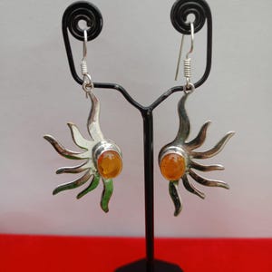 May include: A pair of silver dangle earrings featuring a sunburst design with curved rays extending from a central, oval-shaped orange amber stone. The earrings hang from silver hooks and have a polished metallic finish with a warm amber center.