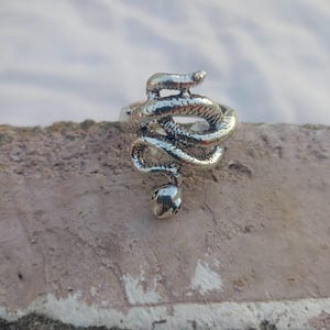 Könnte beinhalten: Ein silberfarbener Ring in Form einer aufgerollten Schlange. Der Körper der Schlange umschlingt den Finger, wobei Kopf und Schwanz die Enden des Rings bilden. Der Ring hat eine detaillierte, strukturierte Oberfläche, vor neutralem Hintergrund.