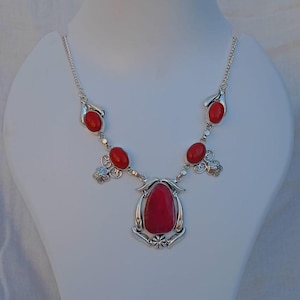 Op de afbeelding: Een zilveren ketting met rode ovale en druppelvormige stenen. De ketting heeft een grote, centrale rode steen en kleinere stenen langs de ketting. Het ontwerp bevat decoratieve zilveren accenten.