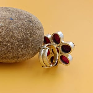 Op de afbeelding: Een zilveren ring met een bloemvormig ontwerp met zes ovale, donkerrode stenen gezet in de metalen bloemblaadjes. De ring heeft een gladde, verstelbare zilveren band en is geplaatst naast een gladde, ronde, beige riviersteen.