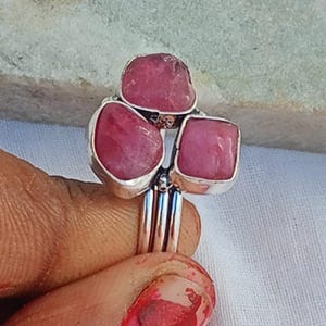 Puede incluir: Un anillo de plata con tres piedras preciosas rosas. Las piedras preciosas tienen forma irregular y están engastadas en una banda de plata. El anillo es una joya única.