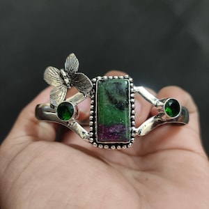 May include: Silver bracelet with a central rectangular green and red gemstone, two green gemstones, and a butterfly detail. The open-design bracelet is held in a hand. The gemstones are set in a silver metal.