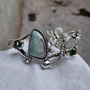 Könnte beinhalten: Ein silberfarbenes Armband mit einem großen, hellblauen und roten Edelstein in der Mitte. Das Armband ist mit zwei grünen Edelsteinen und dekorativen Schmetterlings- und Blumenakzenten verziert. Das Armband liegt auf einer weißen Oberfläche.
