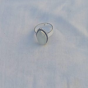 Puede incluir: Un anillo de plata con una piedra grande, ovalada y verde pálido. La piedra está rodeada por un borde de cuentas. La banda del anillo es lisa y brillante. El anillo está sobre una superficie blanca.