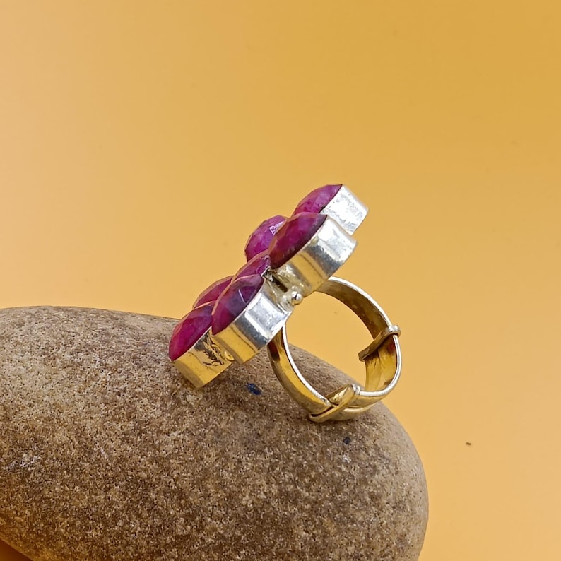 Op de afbeelding: Een zilveren ring met een cluster van gefacetteerde, donkerroze, ronde stenen in een bloemmotief. De ring heeft een verstelbare, open band met een metallic afwerking, waarbij de stenen vanuit een zijprofiel worden getoond.