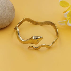 May include: A gold-toned metal bracelet shaped like a coiled snake. The design features a smooth, polished snake head at one end and a slender, undulating body that forms an open-ended circular band. The metal has a reflective, metallic finish.
