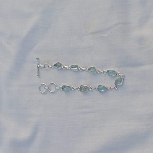 May include: A silver bracelet featuring light blue, irregularly shaped gemstones. The gemstones are linked together with a silver chain and a toggle clasp. The bracelet is displayed on a white, textured surface.