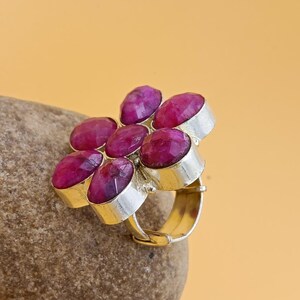 Op de afbeelding: Een zilveren ring met een cluster van zes gefacetteerde, diep roze-rode robijnen in een bloemvormig patroon. De stenen zijn gezet in individuele zilveren zettingen en de ringband is gemaakt van glad, gepolijst zilverkleurig metaal.