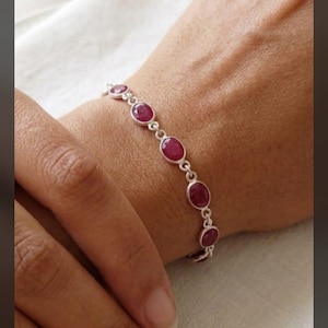 May include: A silver chain bracelet featuring a series of oval-shaped, deep pinkish-red gemstones set in individual metal bezels. The bracelet is composed of multiple linked segments, each holding a single smooth, polished stone in a consistent pattern.