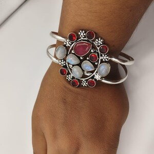 Può includere: Un bracciale a fascia in argento con un elemento centrale circolare decorato con diverse pietre di luna a goccia e gemme rotonde sfaccettate di colore rosso intenso. La struttura in metallo presenta piccoli accenti floreali tra le pietre, creando un design dettagliato.