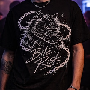 May include: Black t-shirt featuring a detailed white graphic of a wolf's head wearing a muzzle, encircled by a chain design. The text "Bite Risk" is written in a stylized font below the wolf illustration. The shirt is a dark color.