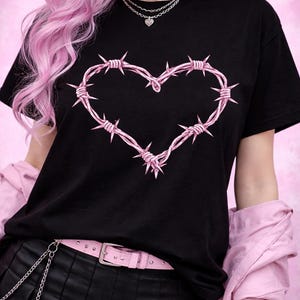 以下が含まれることがあります： ピンクの有刺鉄線ハートのデザインが特徴の黒いTシャツ。モデルは黒いチョーカー、重ね着のネックレス、ピンクのベルト、黒いプリーツスカートを着用しています。全体的な美学はエッジの効いたオルタナティブです。
