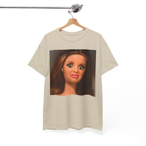 Könnte beinhalten: Ein beiges T-Shirt mit kurzen Ärmeln und einem großen quadratischen Grafikdruck auf der Brust. Der Druck zeigt ein Nahporträt einer Puppe mit langem braunem Haar, weit aufgerissenen Augen und gespitzten rosa Lippen vor dunklem Hintergrund.