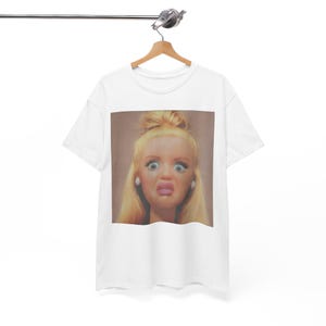 May include: A white short-sleeved t-shirt featuring a square graphic print on the chest. The print displays a close-up portrait of a blonde doll with a high bun hairstyle, wide blue eyes, and a surprised facial expression with pink lips.