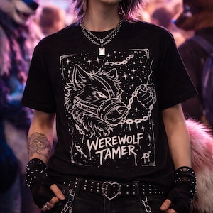 May include: Black t-shirt with a white graphic of a wolf wearing a muzzle and held by a chain. The text "WEREWOLF TAMER" is below the graphic. The shirt is worn with a silver necklace and black studded gloves.