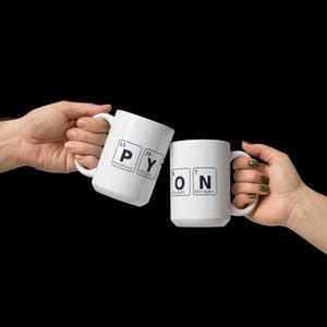以下が含まれることがあります： 黒い文字と周期表の元素が描かれた、2つの白いセラミックマグカップ。1つのマグカップには「PY」とリンとイットリウム、もう1つには「ON」と酸素と窒素が表示されています。マグカップは黒い背景に対して持ち上げられています。