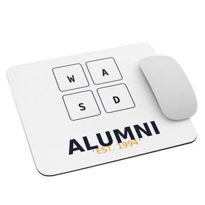 May include: White rectangular mousepad with the letters W, A, S, and D in a grid, and the word "ALUMNI" with "EST. 1994" below it. A white wireless mouse is positioned to the right of the mousepad.