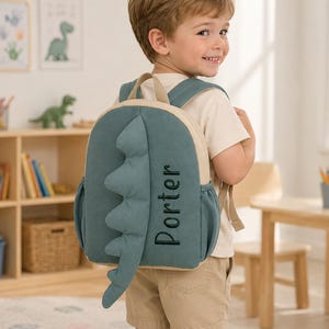 May include: A teal dinosaur-themed backpack featuring a plush, jagged spine detail down the center and the name Porter embroidered in dark green thread on the side. The bag includes adjustable shoulder straps and side pockets for storage.