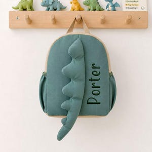 Puede incluir: Una mochila de color verde azulado con una fila vertical de púas de dinosaurio acolchadas en el centro y el nombre Porter bordado en hilo verde oscuro. La mochila tiene bolsillos laterales y cuelga de un perchero de madera clara con cinco ganchos.