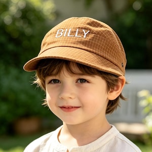 May include: A brown baseball cap featuring a subtle grid pattern and the name BILLY embroidered in white capital letters on the front panel. The cap has a curved brim and a structured crown, providing a classic look with a clean, simple design.