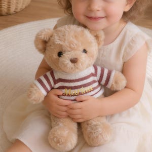 Puede incluir: Una niña con un vestido color crema sostiene un oso de peluche. El oso tiene un suéter a rayas marrones y blancas con la palabra "Meertime" bordada. El fondo incluye una cesta de mimbre y un jarrón con flores.