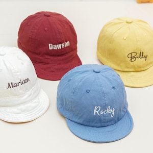 May include: Four baseball caps in white, burgundy, yellow, and blue. Each cap has a curved peak and embroidered name: "Mariam", "Dawson", "Billy", and "Rocky". The caps are made of a soft fabric.