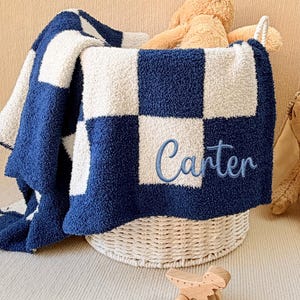 May include: A navy blue and white checkered baby blanket with the name "Carter" embroidered in blue. The blanket is folded in a white wicker basket, with a stuffed bunny and wooden toy nearby. The blanket is soft and plush.
