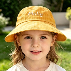 May include: A mustard yellow bucket hat featuring a subtle grid pattern and the name Molly embroidered in white script lettering on the front. The hat has a wide, downward-sloping brim and a rounded crown, constructed from a lightweight fabric.