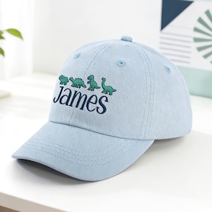 Könnte beinhalten: Hellblaue Baseballkappe mit dem Namen "James" in Dunkelblau gestickt, zusammen mit grünen Dinosaurier-Illustrationen. Die Kappe hat einen gebogenen Schirm und einen Knopf oben.
