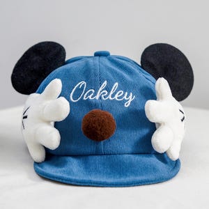 Puede incluir: Gorra de béisbol de pana azul con orejas negras, un pompón marrón y guantes blancos. El nombre "Oakley" está bordado en la parte delantera. La gorra está diseñada para parecerse a un popular personaje de dibujos animados.