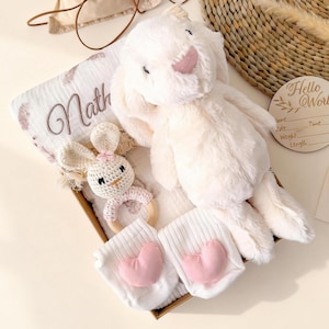 Op de afbeelding: Een baby cadeau set in een doos, met een pluche witte konijn, een gepersonaliseerde deken met de naam "Math", een gehaakte konijn rammelaar en witte sokken met roze hart accenten. Een houten schijf met "Hello World" is ook inbegrepen.