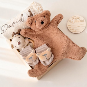 Peut inclure: Un coffret cadeau pour bébé dans une boîte en carton. L'ensemble comprend une couverture ours en peluche marron, une couverture beige avec le nom "David", un hochet en forme de cerf et deux paires de chaussettes à tête d'ours. Un disque en bois avec "Hello World" est également inclus.