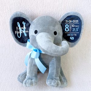 Op de afbeelding: Een grijze pluche olifant met een blauw lint om de nek. De oren van de olifant hebben donkerblauwe accenten met witte tekst. Op één oor staat "Noah Melissa" en op het andere "11-26-2025, 3,6 kg, 54,6 cm, 20:52 uur."