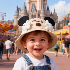 Puede incluir: Un sombrero de pescador beige con orejas negras de Mickey Mouse y guantes blancos. El sombrero tiene bordado el nombre "Charli". El fondo muestra un castillo y atracciones.
