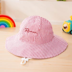 May include: A bucket hat featuring a red and white vertical striped pattern. The front of the hat is embroidered with the name Ramon in a dark red script font. A white adjustable chin strap with a plastic toggle is attached to the brim.
