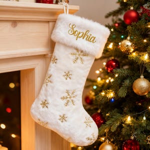 May include: A white Christmas stocking with gold embroidered snowflakes and the name "Sophia" in gold script. The stocking has a fluffy white cuff and is hanging near a decorated Christmas tree.