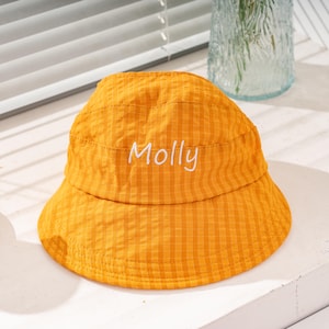 Könnte beinhalten: Ein leuchtend gelber Fischerhut mit einem dezenten Karomuster auf dem gesamten Stoff. Das Wort Molly ist in weißer Schreibschrift mittig auf der Vorderseite der Krone eingestickt. Der Hut hat eine abgerundete Oberseite und eine schräge Krempe.
