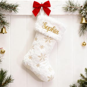 May include: A white Christmas stocking with gold snowflake embroidery and a faux fur cuff. The name "Sophia" is embroidered in gold on the cuff. A red bow hangs above the stocking, with festive greenery and gold bells in the background.