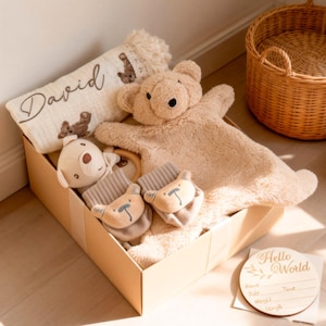 Peut inclure: Un coffret cadeau pour bébé beige dans une boîte en carton. L'ensemble comprend une couverture en peluche en forme d'ours, un petit hochet ours, deux moufles en forme d'ours et une couverture personnalisée avec le nom "David". Un panneau en bois dit "Hello World".