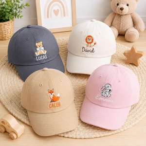 May include: Four baseball caps in charcoal grey, cream, tan, and light pink. Each cap features an embroidered animal and a name: a bear labelled LUCAS, a lion labelled David, a fox labelled CALEB, and a rabbit labelled Emma, all with stitched details.