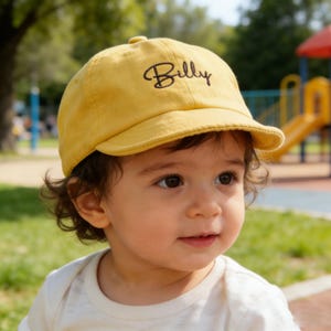 Puede incluir: Gorra de béisbol de color amarillo mostaza con el nombre "Billy" bordado en escritura negra. La gorra tiene una visera curva y la lleva un niño. El fondo muestra un parque con hierba verde y juegos.